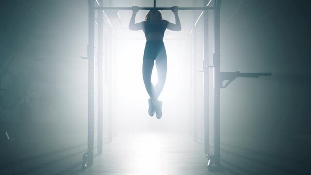 Silhouette of a sporty woman lifting her body while exercising at the gym studio. Female athlete hanging from a bar by her hands and pulling herself upward. High quality 4k footage