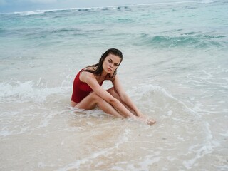 A woman in a red swimsuit sits on the sand by the ocean in the waves at Bali Island Beach and looks...