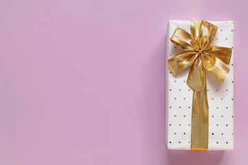 Gift box with golden ribbon and bow on a light purple background. Top view. Copy space.