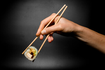 female hand accurate holds unagi sushi roll with smoked eel with chopsticks on dark background