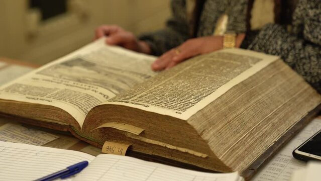 Librarian turns pages of old large-sized book looking for information and holding fingers along lines at table in library closeup