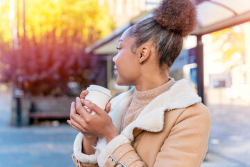 Cheerful African woman holds cup of coffee. A smiling curly brunette lady in brown sweater waiting a tram.