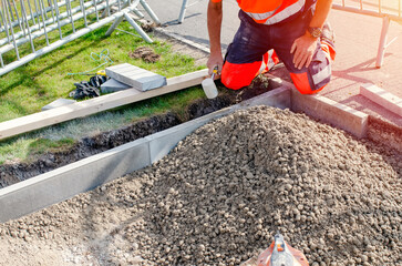 Builder in orange ho-viz protective closing placing edging pin kerb into semi-dry concrete using a string line to keep them straight during construction of the footpath