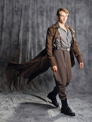 hero in a military style with a gun in his hands, a young man in a long brown coat and breeches with suspenders. Photo in the studio on a gray background