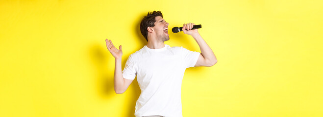 Young man singer holding microphone, reaching high note and singing karaoke, standing over yellow...
