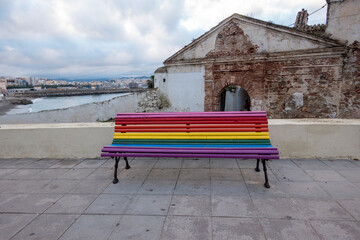City of Ceuta, Spain. A bench in the street with the colors of the rainbow and a dilapidated house...