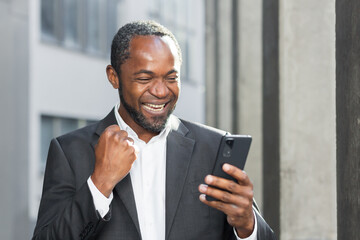 Successful african american boss outside office building using phone businessman in suit celebrating victory, successful achievement, man holding hand up triumph gesture, reading online notification - Powered by Adobe