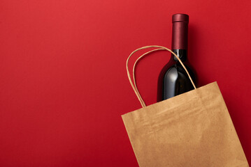 Paper bag with dark glass bottle of wine, alcohol present. Flat lay on maroon background, zero...