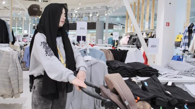 Muslim woman customer with child in baby carriage enjoys walking in clothes department. Mother wants to find perfect match and make purchase