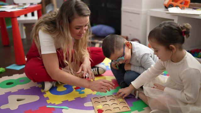 Teacher with boy and girl playing with maths puzzle game sitting on floor at kindergarten