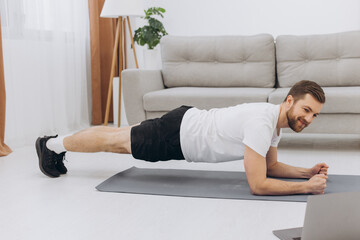 Training At Home. Sporty man doing yoga plank while watching online tutorial on laptop, exercising in living room, free space