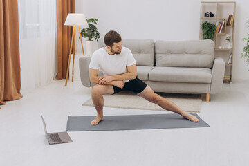 Positive athletic bearded middle-aged man stretching at home, using laptop, watching sport videos on Internet, having fitness class online, copy space. Healthy lifestyle, sport on self-isolation