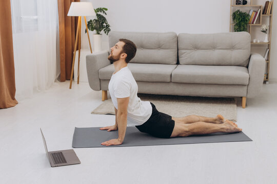 Positive athletic bearded middle-aged man stretching at home, using laptop, watching sport videos on Internet, having fitness class online, copy space. Healthy lifestyle, sport on self-isolation