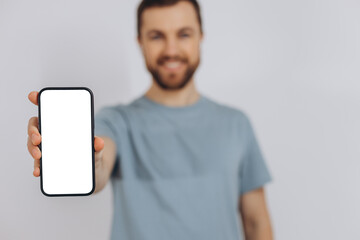 Fototapeta na wymiar Cheerful Emotional Bearded Man Holding Blank Cell Phone In Hand Showing White Screen To Camera Pointing At You, Happy Millennial Guy Recommending New Application Or Mobile Website, Mockup Banner