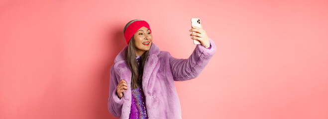 Fashion concept. Beautiful asian senior lady taking selfie on smartphone, posing in party...