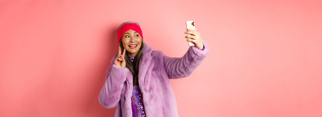Fashion concept. Cool and cheerful asian senior woman taking selfie with peace sign, wearing trendy...