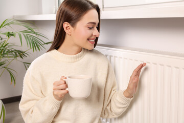 Woman holding cup with hot drink and warming hand on heating radiator indoors