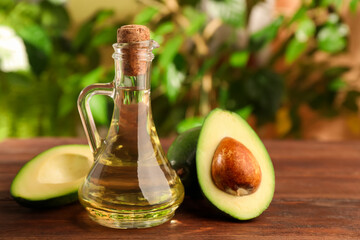 Glass jug of cooking oil and fresh avocados on wooden table against blurred green background
