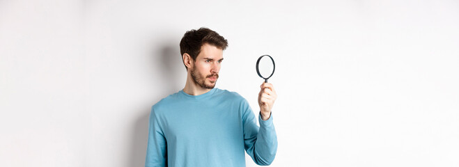 Young handsome man look through magnifying glass with curious face, investigating or searching for...