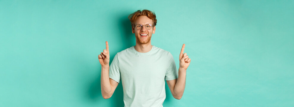 Image of handsome bearded man with red hair, wearing t-shirt and glasses, smiling happy and pointing fingers up, showing promo offer, standing over turquoise background