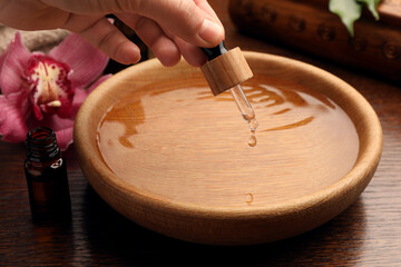 Woman dripping essential oil into bowl at wooden table, closeup. Aromatherapy treatment