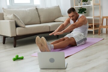 Man sports, watching tape of workout on phone and repeating exercises sports blogger with laptop training online, pumped up man fitness trainer works out at home, the concept of health and body beauty