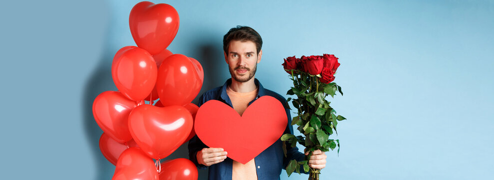 Valentines Day Romance. Young Man With Bouquet Of Red Roses And Heart Balloons Smiling, Bring Presents For Lover On Valentine Date, Standing Over Blue Background