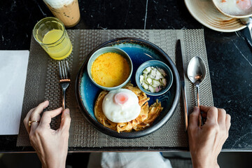 Breakfast - Roti with chicken green curry, fried egg, and cucumber in a sweet sauce served on blue plate. Hands holding cutlery on the table, preparing to have a breakfast