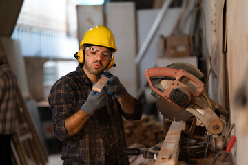 Young white carpenter man blowing dust on wood plank after cutting from a circular saw machine in...