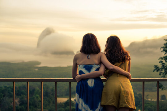 Mother and daughter travel together, standing at view point to see sunrise with small island in ocean view background at resort. Travelers hug together with love, family relationship. Journey concept 