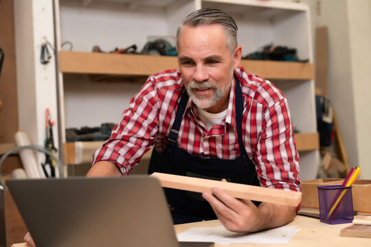 Elderly carpenter man checking wood type on laptop before order wooden to build crafts handmade furniture. Business carpentry shop owner pay attention to details in furnish assembly. Joiner concept. 