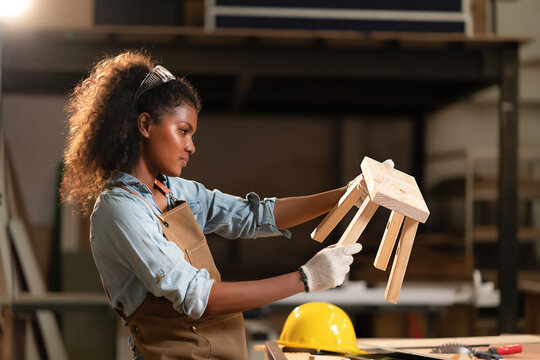 Young black carpenter woman wearing gloves hands, holding small wooden chair in hand after manual assembly. Handmade furniture building based on custom design, crafts joiner maker concept.
