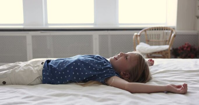 Side view cute little 6s girl take break, falling on bed with arms raised enjoy rest lying on fresh white bedsheets smile relaxing alone at home. Comfortable mattress for healthy sleep, fatigue relief