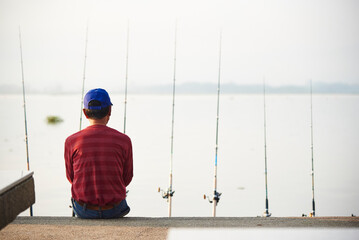 Back of a man sitting with many fishing rods by the lake with blurry land and white sky in the...