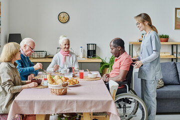 Senior people sitting at table and eating with caregiver helping man with disability in nursing home