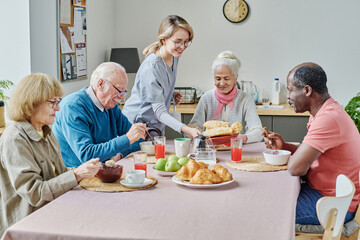 Young caregiver serving senior people at table, they sitting at table and eating food and she setting the table