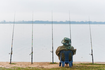 Back of a relaxing man sitting in a small chair with many fishing rods by the lake with blurry land and white sky in the background