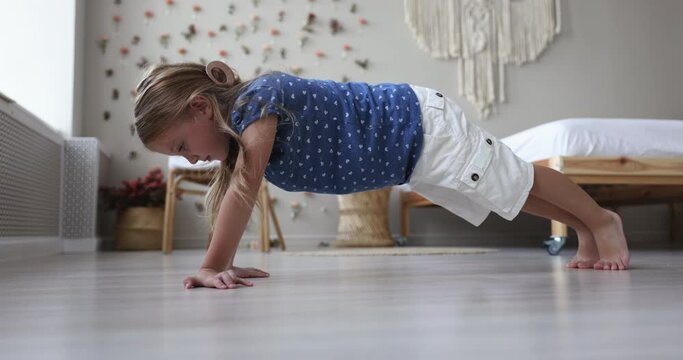 Cute little girl in casual clothes exercising alone in modern warm bedroom, getting physically stronger stands in a horizontal position doing plank, full length, side view. Kid practicing yoga at home