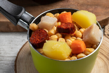 Cooked (Cocido, Guiso) stew, chickpeas with chorizo, bacon, carrot and potato in a green saucepan and on wooden board. Concept of traditional Spanish cuisine.