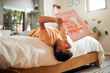 Happy child reading book on bed for home learning, language education and creative development upside down. Smart, intelligent and Indian kid with English story for fun, relax and holiday activity - Powered by Adobe