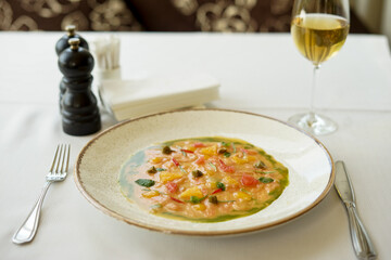salmon carpaccio in a white plate on the table with a glass of white wine
