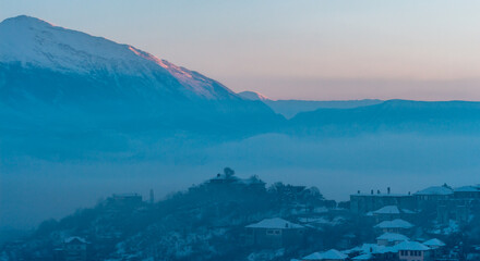 Winter landscape, covered with snow. Panorama of Gjirokastër (Gjirokaster), Albania, with the gorgeous Mali i Gjerë mountains that surround the city. Morrning light with mist. Blue hour.