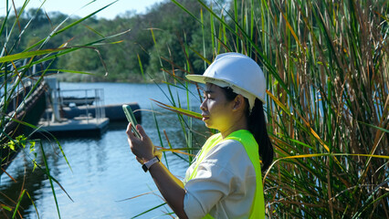 Environmental engineer wearing a white helmet uses a mobile phone to record data analyzing oxygen levels in a reservoir. Water and ecology concept.