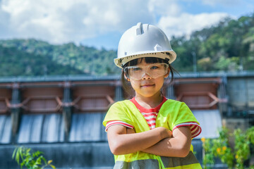Portrait of a little girl engineer wearing a green vest and white helmet smiling happily on the background of the dam. Concepts of environmental engineering, renewable energy and love of nature.