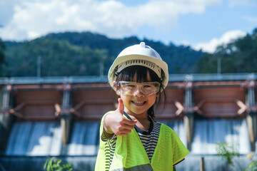 Portrait of a little girl engineer wearing a green vest and white helmet smiling happily on the background of the dam. Concepts of environmental engineering, renewable energy and love of nature.