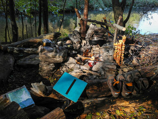 Campfire with pots and metal utensils by the river, a bunch of mushrooms by the fire, hiking boots and a topographic map
