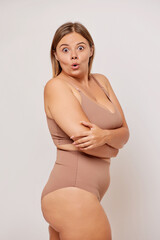 Obraz premium Side view of amazed brunette curvy women in beige lingerie poses half turned to the camera, embraced her natural body, isolated over white wall