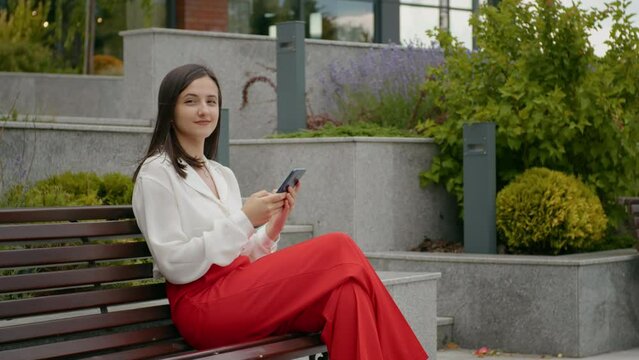 Business woman working outside office on a bench looking at hte camera. Technology addicted female student having a break outside university building. Technology era.