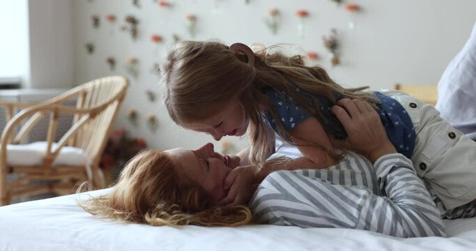 Close up side view loving little girl kisses mothers nose, affectionate mom tickling daughter, resting lying on bed in bedroom. Enjoy moment of caress, warmth and tenderness feeling unconditional love
