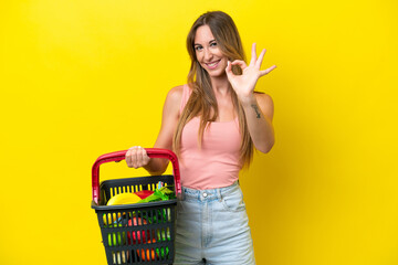 Young caucasian woman holding a shopping basket full of food isolated on yellow background showing ok sign with fingers